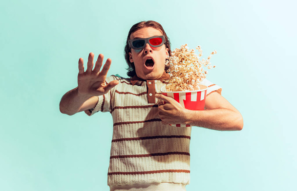 Portrait of young man posing with popcorn basket isolated over light blue studio background. Looks super excited and shocked. Concept of youth, fashion, lifestyle, emotions. Copy space for ad - Φωτογραφία, εικόνα