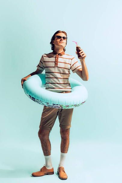 Portrait of young cheerful man in casual outfit and swimming circle, drinking coke, posing isolated over light blue studio background. Concept of youth, fashion, lifestyle, emotions. Copy space for ad - Foto, afbeelding