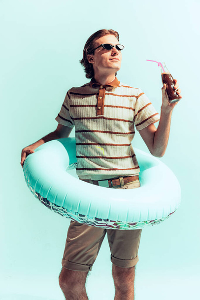 Портрет молодого веселого человека в повседневной одежде и кругу для купания, пьющего кокаин, позирующего изолированно на светло-голубом фоне студии. Концепция молодости, моды, образа жизни, эмоций. Копирование места для рекламы - Фото, изображение