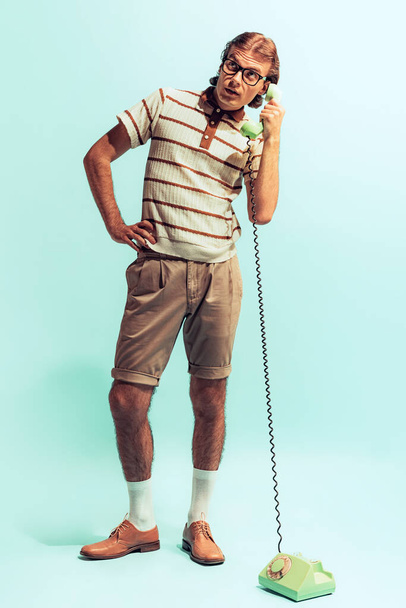 Portrait of young emotive man in retro style outfit posing with old-fashioned phone isolated over light blue studio background. Concept of youth, fashion, lifestyle, emotions. Copy space for ad - Fotoğraf, Görsel