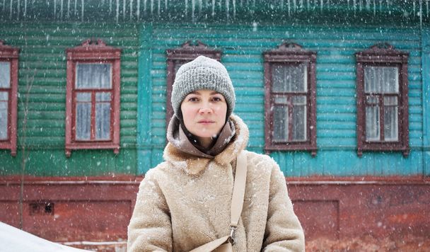 Młoda kobieta stoi w ciepłym futrze. Strzał z przodu patrząc w kamerę. W tle zielony drewniany budynek w Czernihowie, Ukraina. śnieżna zima - Zdjęcie, obraz