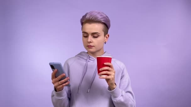 Модна молода жінка покоління Z з прохолодною короткою зачіскою, використовуючи мобільний телефон, п'є одноразовий паперовий стаканчик, розважаючий Інтернет, інтернет-покупки. Ізольований студійний знімок на фіолетовому фоні
 - Кадри, відео