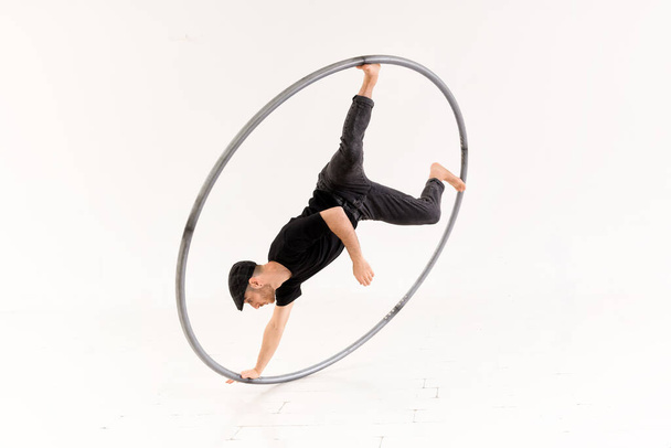 Full body of active barefoot acrobat performing one arm coin stunt with cyr wheel against white background in light studio - Photo, Image