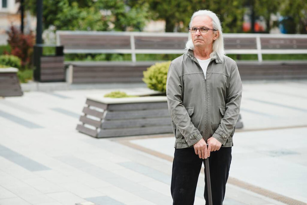 Portrait of senior man holding walking stick while resting in park - Zdjęcie, obraz