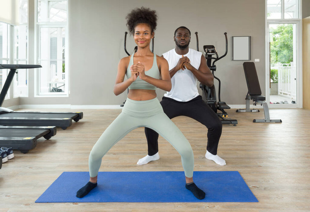 Young curly black hair instructor in sportswear leading exercise class how to do bodyweight squat. Cardio machines are on the background at the gym. - Photo, Image