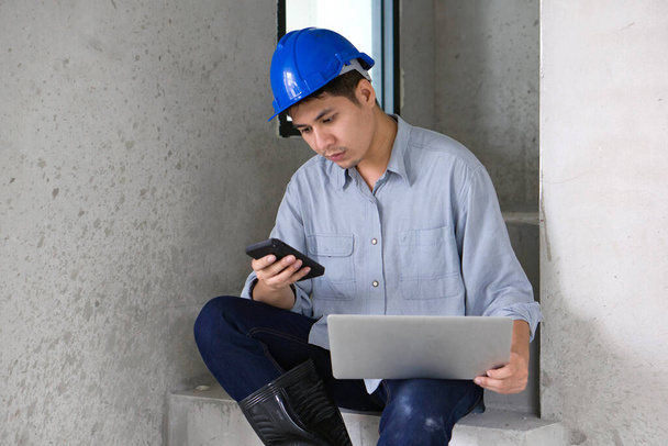 階段に座っている間、携帯電話からラップトップコンピュータにハードハット転送データの若いアジアのエンジニア。住宅事業の工事現場における技術者の作業環境. - 写真・画像