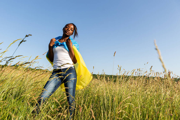 Mujer afroamericana envuelta en ondulaciones de bandera azul amarilla ucraniana ondeando al viento. Símbolo nacional de Ucrania. Apoyar a Ucrania, apoyo internacional y solidaridad - Foto, Imagen