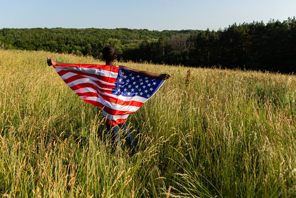 アフリカ系アメリカ人の女性が風になびくアメリカの国旗のはためきに包まれた。7月4日ハッピー!独立記念日だ。星と縞模様。自由の概念. - 写真・画像