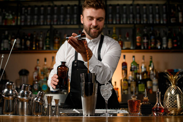 various steel shakers and bottles stand on the bar counter and male bartender masterfully pours an alcoholic drink from jigger in the shaker cup - Foto, Bild