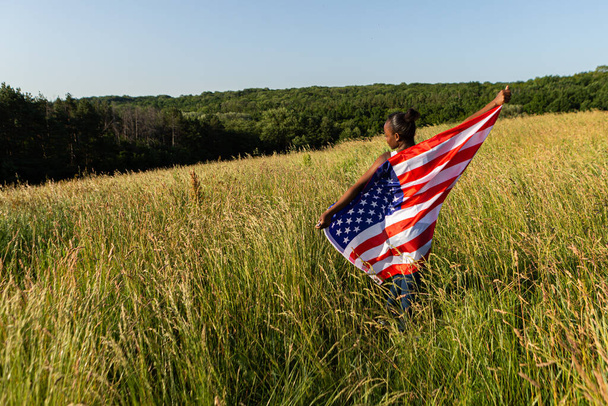 Mulher afro-americana envolta em flutters bandeira americana acenando ao vento. Feliz 4 de Julho! Dia da Independência comemorando. Estrelas e riscas. Conceito de liberdade. - Foto, Imagem
