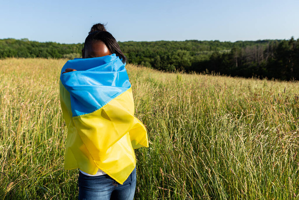 African american woman wrapped in ukrainian yellow blue flag flutters waving in the wind. National symbol of Ukraine. Stand with Ukraine, international support and solidarity - Photo, image