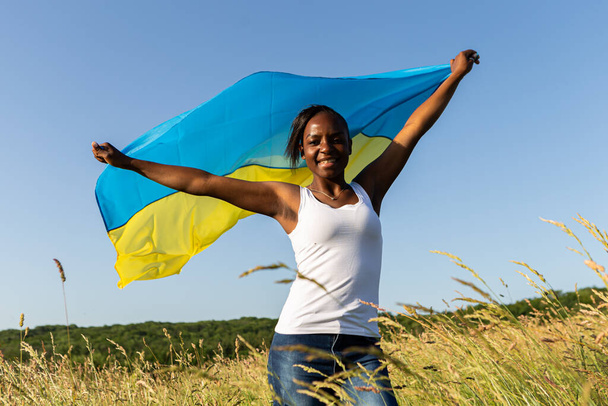 Mujer afroamericana envuelta en ondulaciones de bandera azul amarilla ucraniana ondeando al viento. Símbolo nacional de Ucrania. Apoyar a Ucrania, apoyo internacional y solidaridad - Foto, imagen