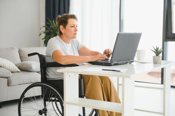 Freelancer in wheelchair using laptop near notebook and papers on table. - Φωτογραφία, εικόνα