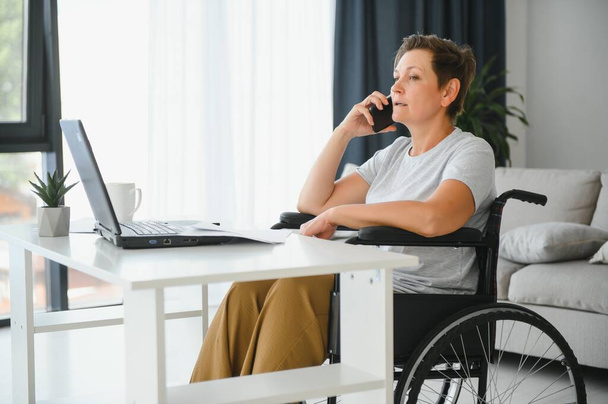 Freelancer in wheelchair using laptop near notebook and papers on table. - Foto, imagen