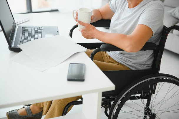Middle age woman using laptop sitting on wheelchair at home. - Foto, afbeelding