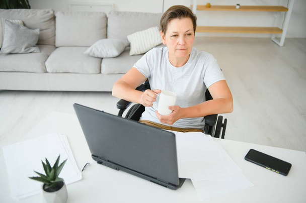 Middle age woman using laptop sitting on wheelchair at home. - Valokuva, kuva