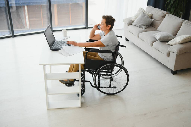 Positive elderly woman in wheelchair working on laptop, IT freelancer, online. - 写真・画像