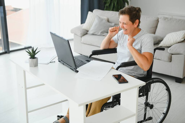 Positive elderly woman in wheelchair working on laptop, IT freelancer, online. - Fotó, kép
