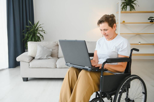 Middle age woman using laptop sitting on wheelchair at home. - Photo, Image