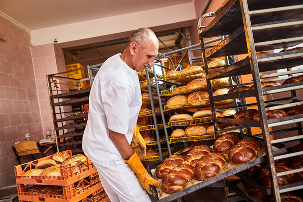 Baker holds a tray with fresh hot bread in his hands against background of shelves with pastries in bakery. Industrial production of bread - Photo, Image
