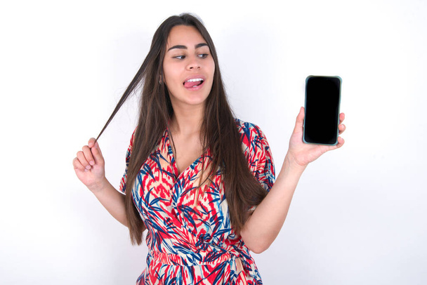 Photo of nice pretty brunette woman wearing colourful dress over white wall demonstrate phone screen hold hair tails - Foto, afbeelding