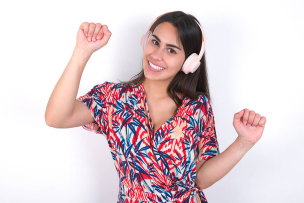 Carefree beautiful brunette woman wearing colourful dress over white background with toothy smile raises arms dances carefree moves with rhythm of music listens music from playlist via headphones - 写真・画像