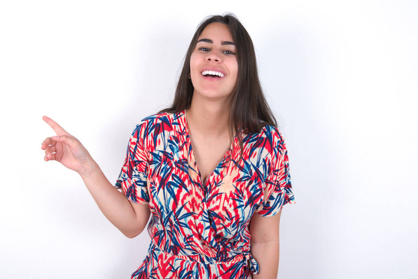 krásná brunetka žena v barevných šatech přes bílou stěnu se šťastně směje body pryč na prázdném prostoru demonstruje nákupní slevu nabídku, vzrušený dobrou zprávou nebo neočekávaný prodej. - Fotografie, Obrázek
