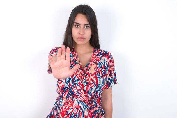 beautiful brunette woman wearing colourful dress over white wall shows stop sign prohibition symbol keeps palm forward to camera with strict expression - Photo, Image