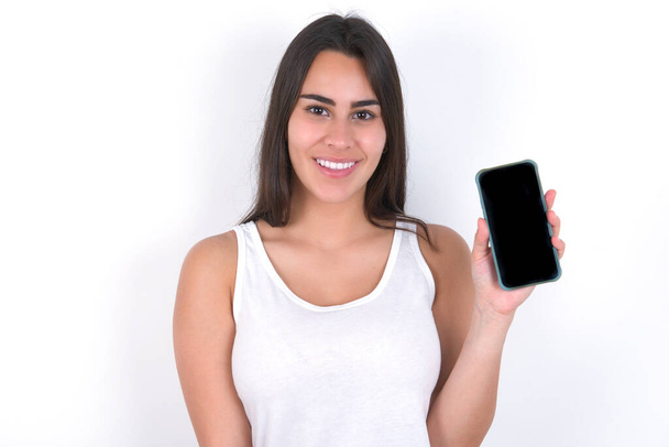 Smiling beautiful brunette woman wearing white top over white wall showing  empty phone screen. Advertisement and communication concept. - Foto, Bild