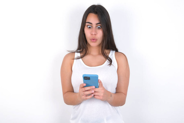 beautiful brunette woman wearing white top over white looks with bugged eyes, holds modern smart phone, receives unexpected message from friend, reads reminder. - Foto, immagini