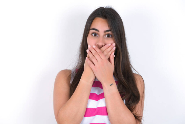 Upset beautiful brunette woman wearing striped crop top over white wall, covering her mouth with both palms to prevent screaming sound, after seeing or hearing something bad. - Photo, Image