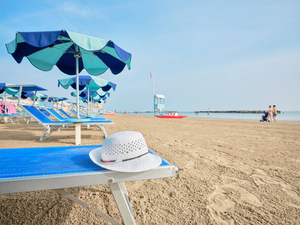 White straw hat on a beach bed with colorful umbrellas on a sunny summer day. In the background a lifeboat alongside a lifeguard tower on the shore of the Adriatic Sea, Italy. - Photo, Image