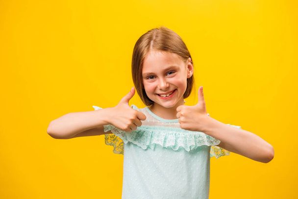 Like and approval. Studio portrait of stylish little children girl gesturing thumbs up and looking at camera. smiling to camera, yellow background - Фото, изображение