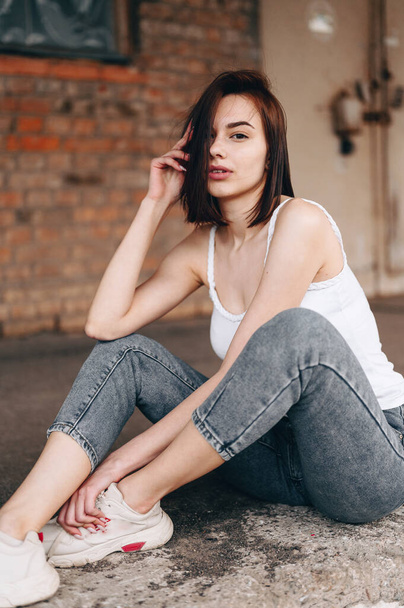 A young woman sits in jeans on the ground in front of an old building. Smart Educated Girl is resting on the street. Leisure. - Фото, зображення