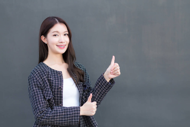 Asian professional working woman who wears black suit with braces on teeth is pointing hand to present as thump up on the dark gray wall. - Photo, Image