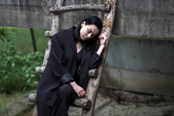 Una bella ragazza ucraina in un abito nero si siede su una scala di legno e cammina attraverso una fabbrica abbandonata. - Foto, immagini