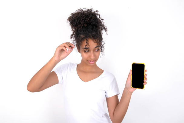 Photo of nice pretty girl demonstrating phone screen, holding hair tails. Young beautiful mixed race woman wearing white t-shirt posing over white wall - Foto, afbeelding