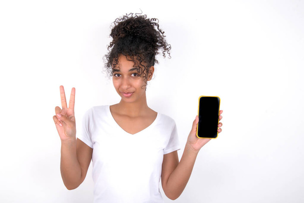 Muchacha sosteniendo un dispositivo moderno mostrando signo v. Joven hermosa mujer de raza mixta con camiseta blanca posando sobre la pared blanca - Foto, Imagen