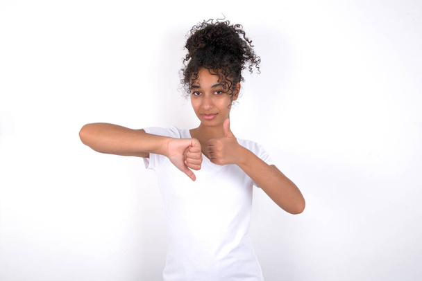 girl showing thumbs up and down sign Young beautiful mixed race woman wearing white t-shirt posing over white wall - Фото, зображення