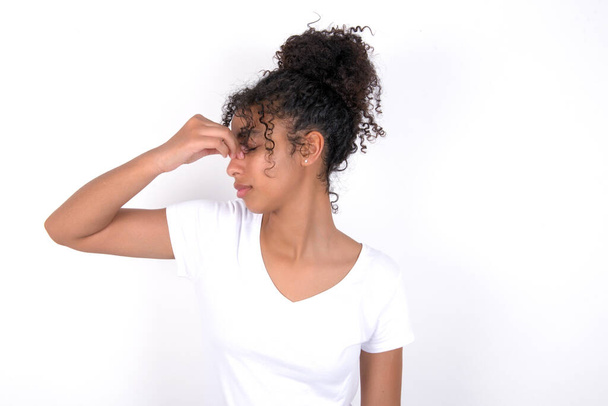 Sad girl suffering from headache holding hand on her face. Young beautiful mixed race woman wearing white t-shirt posing over white wall - Photo, Image