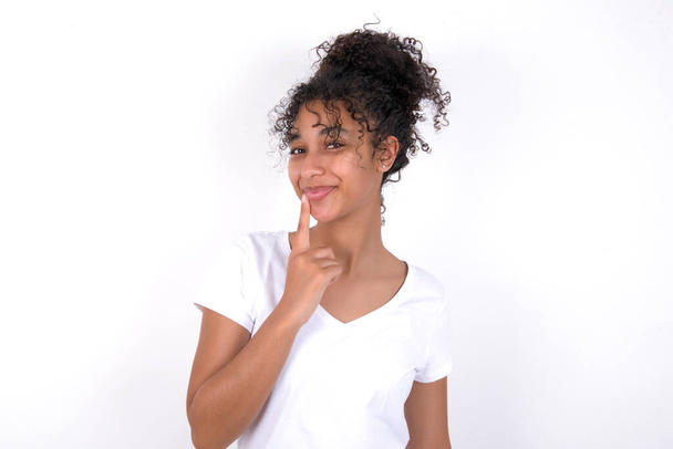 Lovely dreamy girl keeps finger near lips, looks aside copy space. Young beautiful mixed race woman wearing white t-shirt posing over white wall - Photo, Image