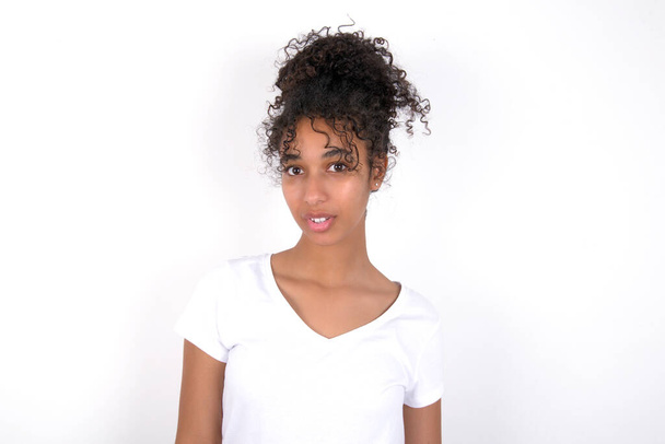 Shocked girl stares bugged eyes keeps mouth opened has surprised expression. Omg concept. Young beautiful mixed race woman wearing white t-shirt posing over white wall - Photo, Image