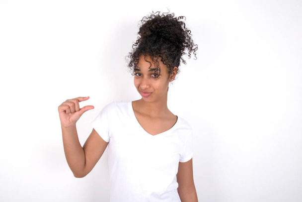Displeased girl shapes little hand sign demonstrates something not very big. Body language concept. Young beautiful mixed race woman wearing white t-shirt posing over white wall - Photo, Image