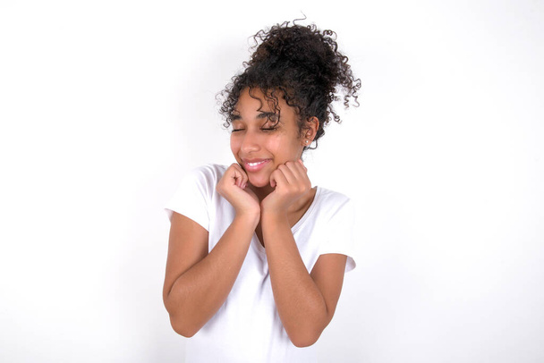 girl grins joyfully, imagines something pleasant, copy space. Pleasant emotions concept. Young beautiful mixed race woman wearing white t-shirt posing over white wall - Photo, Image