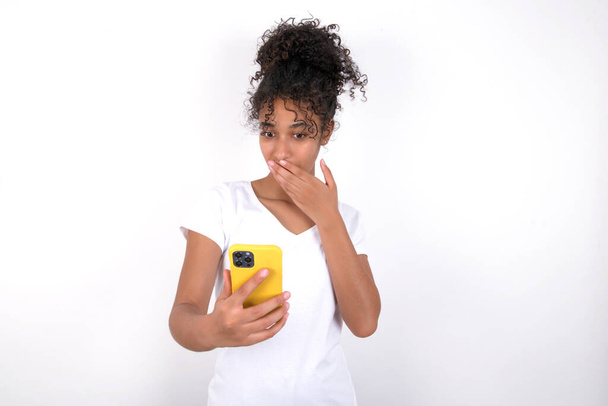 girl being deeply surprised, stares at smartphone display, reads shocking news on website, Omg, its horrible! Young beautiful mixed race woman wearing white t-shirt posing over white wall - Foto, afbeelding