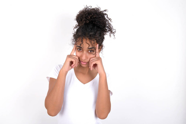 Serious concentrated girl keeps fingers on temples, tries to ease tension, gather with thoughts and remember important information for exam. Young beautiful mixed race woman wearing white t-shirt posing over white wall - Fotó, kép