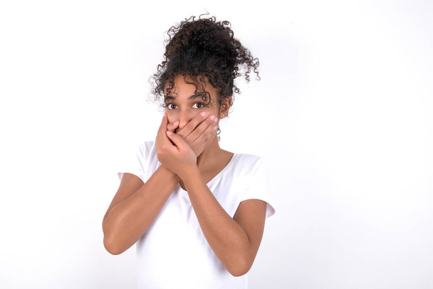 Stunned Young beautiful girl with afro hairstyle wearing white t-shirt over white background covers mouth with both hands being afraid from something or after hearing stunning gossips. - Valokuva, kuva