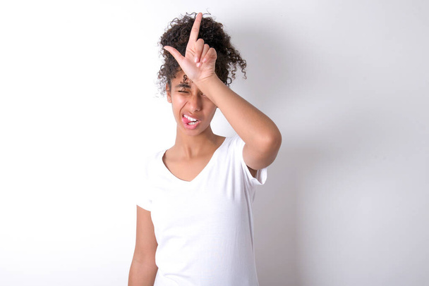 Young beautiful girl with afro hairstyle wearing white t-shirt over white background gestures with finger on forehead makes loser gesture makes fun of people shows tongue - Photo, Image