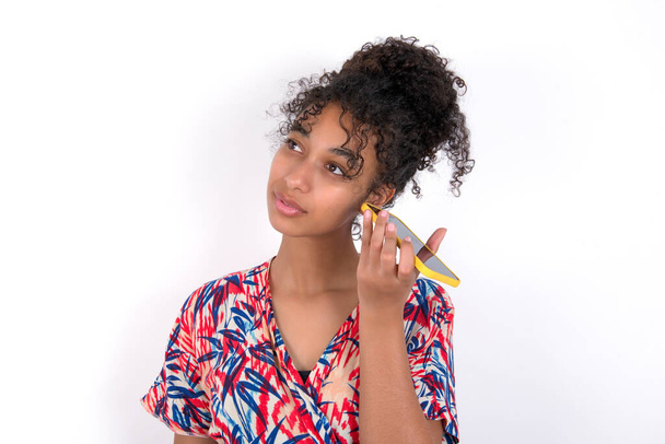 Femme brune souriante portant une robe colorée sur un mur blanc écoutant un message vocal de son smartphone. Concept de communication et technologie. - Photo, image