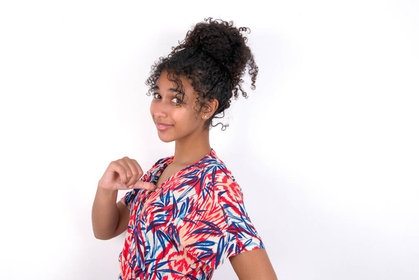 Closeup of cheerful girl looks joyful, satisfied and confident, points at herself with thumb. Young beautiful mixed race woman wearing colourful dress standing over white wall - Photo, Image